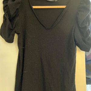 Svart ribbad topp från KappAhl - Snygg svart ribbad topp från KappAhl med v-ringning och korta puffärmar. Perfekt för en stilren look. Passar bra till både jeans och kjol.