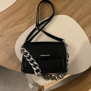 Svart axelväska med kedjedetalj - Svart axelväska från Bershka i krokodilmönstrat lackat fuskskinn med en silverfärgad kedja. Justerbar, avtagbar axelrem. Kedjan går att haka på/av. Stängs med magnetlås. Nyskick!! ✨