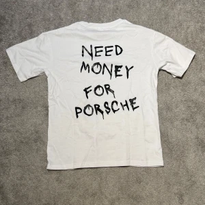 ”NEED MONEY FOR PORSCHE” T-shirt - Vit t-shirt med svart text 'NEED MONEY FOR PORSCHE' på baksidan. Enkel design med rund hals och korta ärmar. Väldigt eftertraktad och populär. Köpt för 559kr. Helt oanvänd i orginalförpackning. Passar 160-175. Tröja med tryck.
