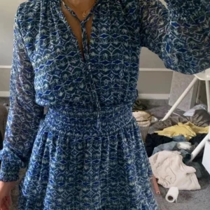 Blå mönstrad klänning - Snygg blå klänning med ett unikt mönster och långa ärmar. Den har en smockad midja som ger en fin passform och ett knytband vid halsen. Perfekt för en stilfull look.