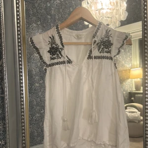 Vit blus med svart broderi - Charmig vit blus med vackert svart blommigt broderi. Blusen har korta ärmar och en knytning.  Ena tofsen har gått upp men det går att fixa med en looband så ser det ut som vanligt🥰