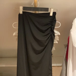 Svart långkjol med rynkdetalj - Elegant svart långkjol med rynkdetalj på sidan. Har en bra smikrande passform om rumpan och en stor slits 