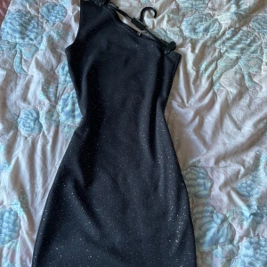 Svart glittrig klänning - Snygg svart klänning med glittrande detaljer. Den har en oneshoulder-design som ger en trendig look. Perfekt för en kväll ute eller en speciell tillställning.