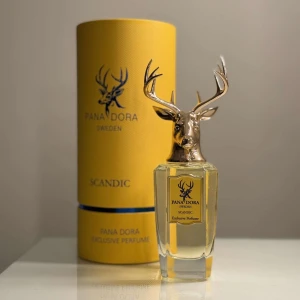Pana Dora Scandic Exclusive Perfume - Elegant parfym från Pana Dora med en unik flaska prydd av ett gyllene hjorthuvud. Flaskan är genomskinlig och cylindrisk, medan förpackningen är gul med blå detaljer. Perfekt för den som söker en exklusiv doftupplevelse.