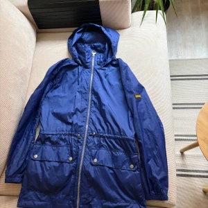 Blå vindjacka från Barbour - Säljer en blå vindjacka från Barbour med huva och dragkedja framtill. Jackan har praktiska fickor med knappar och en justerbar midja för bättre passform.