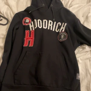 Svart hoodie från Hoodrich - Säljer en svart hoodie från Hoodrich med vita bokstäver och röda detaljer. Hoodien har en stor ficka framtill och justerbar huva med snören. Perfekt för en avslappnad stil.