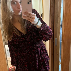 Svart och röd klänning med blommigt mönster - Snygg svart och röd klänning med blommigt mönster och omlott-design. Klänningen har långa ärmar och en smickrande v-ringad hals. Perfekt för en chic look!