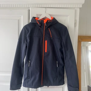 Jacka från Superdry - Jag säljer denna snygga jacka från Superdry. Jackan är av modellen hooded windtrekker och är en softshelljacka som är vind/vattenavvisande. Jackans utsida har en blågrå färg med orangea detaljer. Insidan är täckt med ett bekvämt innerfoder, som har en fin orange färg. Aldrig blivit använd och är därför i mycket fint skick utan defekter. Längst ner har jackan justerbara snören och även vid luvan. Pris kan diskuteras. Tveka inte att fråga om det är någonting du undrar över eller vid fler bilder!☺️