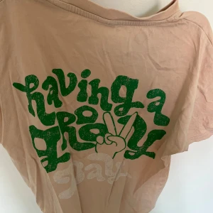 Beige/rosa t-shirt med grönt tryck - Säljer en beige t-shirt med ett coolt grönt tryck som säger 'having a great day' och en peace-symbol. Perfekt för en avslappnad stil. T-shirten har korta ärmar och rund hals.