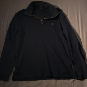 Mörkblå  långärmad tröja från Ralph Lauren - Säljer en stilren mörkblå långärmad tröja från Ralph Lauren med dragkedja vid halsen. Tröjan har en broderad logotyp på bröstet och är perfekt för en laidback look.