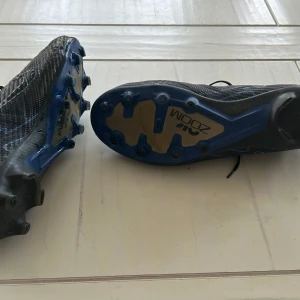 Svarta fotbollsskor från Nike - Säljer ett par svarta Nike fotbollsskor med blå detaljer och snörning. Skorna har en slitstark sula med dobbar för bra grepp på planen. Perfekta för fotbollsträning och matcher. Skick 7/10