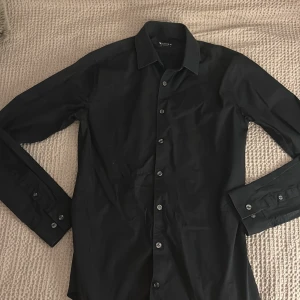 Svart skjorta från Tiger of Sweden - Snygg svart skjorta från Tiger of Sweden med klassisk krage och knappar framtill. Perfekt för en stilren look. Skjortan har långa ärmar och är tillverkad i ett bekvämt material. Storlek S sitter slim