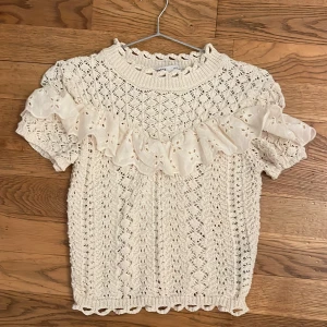 Beige stickad topp från Zara - Säljer en söt beige stickad topp från Zara med vackra spetsdetaljer och volanger. Toppen har korta ärmar och ett fint hålmönster som ger en romantisk känsla. Naturvit färg 🌸
