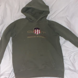 Grön hoodie från GANT - Säljer en grön hoodie från GANT med en stor ficka framtill och broderad logga på bröstet. Perfekt för en avslappnad stil. Huvan ger extra mysfaktor.