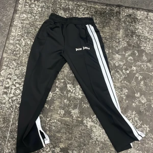 Svarta träningsbyxor från Palm Angels - Snygga svarta träningsbyxor från Palm Angels med vita sidoremsor och dragkedja vid bensluten. Perfekta för en sportig look med en touch av stil.