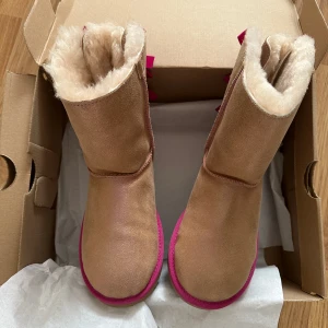Bruna boots med rosa detaljer från UGG - Säljer ett par bruna UGG boots med fluffigt foder och rosa detaljer. De har dekorativa rosa rosetter på baksidan och en rosa sula. Perfekta för att hålla fötterna varma och stiliga under kallare dagar.