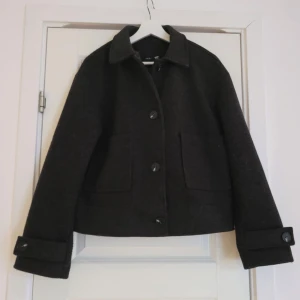 Svart ulljacka med knappar - Snygg svart ulljacka med knappar framtill och klassisk krage. Jackan har en kortare passform och långa ärmar med knappar vid ärmsluten. Perfekt för kyligare dagar!