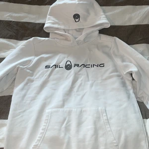 Vit hoodie från Sail Racing - Säljer en stilren vit hoodie från Sail Racing med logga på bröstet och huvan. Tröjan har en klassisk känguruficka och långa ärmar. Perfekt för en avslappnad stil.