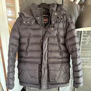 Svart dunjacka från Colmar - Säljer en snygg svart dunjacka från Colmar. Jackan har en avtagbar huva och praktiska dragkedjor på fickorna. Perfekt för kyliga dagar med sin vadderade design och stilrena look. Passar både till vardags och mer uppklädda tillfällen. Jackan har även 6 fickor på framsidan och en stor innerficka! Fickorna få bröstet har fleece i fickorna som håller dina händer varma!