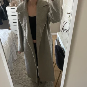 Beige/grå kappa - En stilren beige/grå kappa med lång passform och krage. Kappan har långa ärmar och knäppning framtill, även ett knytband! 