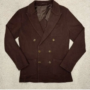 Brun kavaj från John Henric - Snygg brun kavaj från John Henric med dubbelknäppning och sex knappar framtill. Kavajen har en klassisk design med breda slag och är tillverkad i ett mjukt material. Perfekt för en stilren look.