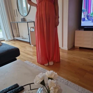 Korallfärgad långklänning - Elegant korallfärgad långklänning med omlott-design och v-ringad hals. Perfekt för sommarens festligheter. Klänningen har en smickrande passform och är ärmlös, vilket gör den sval och bekväm.