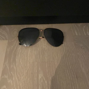 Svarta pilotbågar solglasögon med guldfärgade detaljer från Daniel Wellington - Snygga solglasögon i klassisk pilotmodell med svarta glas och tunna guldfärgade metallbågar. Skalmarna har svarta ändar för extra komfort. Perfekt accessoar för en stilren look.