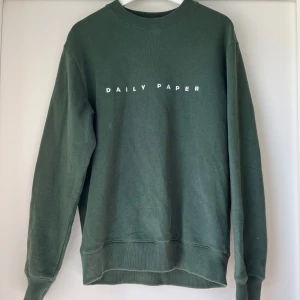 Mörkgrön sweatshirt från Daily Paper - Mörkgrön sweatshirt från Daily Paper med vit logotyp på bröstet. Endast använd en gång! Nypris 1200 kr. Tröjan har rund halsringning, ribbade muddar vid ärmslut och nederkant samt lång ärm. Perfekt för en avslappnad men stilren stil. 100% bomull. 