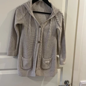 Beige stickad kofta med huva från Newbie - Mysig beige stickad kofta från Newbie med huva, två fickor framtill och fina flätade detaljer längs ärmar och framkant. Perfekt för lager-på-lager under kyliga dagar.