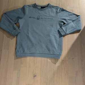 Ljusblå sweatshirt från Sail Racing - Säljer en ljusblå sweatshirt från Sail Racing med tryckt logga på bröstet. Tröjan har rund halsringning och långa ärmar. Perfekt för en avslappnad stil och enkel att matcha med jeans eller shorts.
