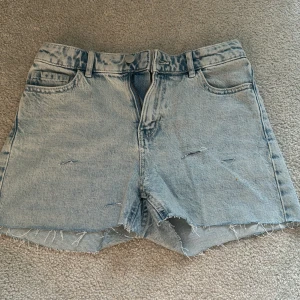 Ljusblå jeansshorts från Lindex - Ljusblå jeansshorts från Lindex med råa, fransiga benslut och slitna detaljer framtill. Klassisk femficksmodell med bälteshällor och knappgylf. Märkt med 'DETROIT' baktill på linningen.
