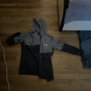 Grå hoodiejacka från Under Armour - Säljer en grå och mörkgrå hoodiejacka från Under Armour med dragkedja och huva. Jackan har lång ärm och en sportig look, perfekt för träning eller chill. Klassisk logga på bröstet och bekväm passform.