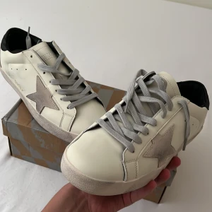 Vita sneakers med grå stjärna från Golden Goose - Säljer ett par vita sneakers från Golden Goose med grå stjärndetalj på sidan och grå snörning. Skorna har en platt sula och svart detalj vid hälen. Perfekta för en avslappnad och trendig stil.