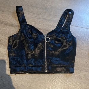 Svart croppad topp med dragkedja från Divided - Säljer en svart croppad topp från Divided i glansigt material. Toppen har breda axelband och en dekorativ dragkedja framtill med en rund detalj. Perfekt för att lyfta en outfit med lite extra edge.