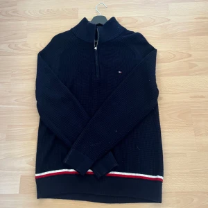 Marinblå stickad tröja med dragkedja från Tommy Hilfiger storlek M - Säljer en marinblå stickad tröja från Tommy Hilfiger med halv dragkedja och diskret logga på bröstet. Tröjan har långa ärmar och röd-vit rand vid nederkanten. Perfekt för kyliga dagar.