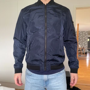 Bomberjacka - Mörkblå tunnare bomberjacka från Jack & Jones Core i strl M. Dragkedja, fickor i fram och svart mudd på ärmar och nedre kant. Använd få gånger