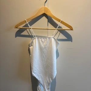 Vit ärmlös bodysuit med tunna axelband - Säljer en enkel och stilren vit bodysuit med tunna axelband. Perfekt att matcha med jeans eller kjol för en clean look. Materialet känns mjukt och stretchigt.