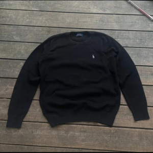Svart stickad tröja från Polo Ralph Lauren - Klassisk svart stickad tröja från Polo Ralph Lauren med rund halsringning och diskret broderad logga på bröstet. Perfekt för en stilren och enkel look. Långa ärmar och ribbade muddar.