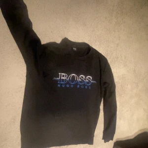 Svart hoodie utan luva från Hugo Boss - Svart sweatshirt från Hugo Boss med stort vitt och blått BOSS-tryck på bröstet. Klassisk rund halsringning och långa ärmar. Perfekt för en stilren och avslappnad look. ÄKTA INTE ANVÄND!