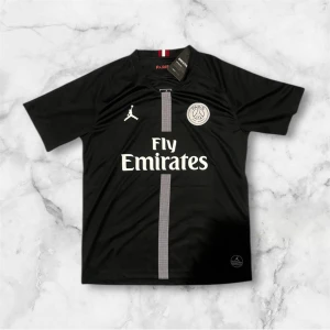 Svart PSG fotbollströja Fly Emirates - Säljer en svart PSG fotbollströja med Fly Emirates-tryck på bröstet. Tröjan har korta ärmar, vit logga och emblem samt en grå rand längs mitten. Tillverkad i lätt och ventilerande material, perfekt för fotbollsträning eller match.