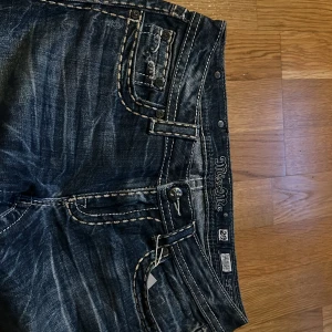 Blå jeansbyxor från Miss Me med kontrastsömmar - Snygga blå jeansbyxor från Miss Me med tydliga kontrastsömmar och dekorativa detaljer på bakfickorna. Byxorna har klassisk femficksdesign, normal passform och bootcut-stil. Perfekta för dig som gillar jeans med lite extra detaljer.