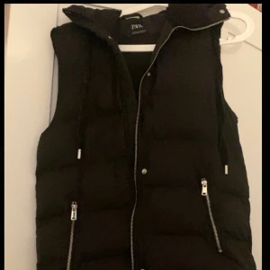 Svart väst från ZARA - Säljer en svart dunväst från ZARA med huva och dragkedja framtill. Västen har två fickor med dragkedjor och en hög krage. Den är som ny, storlek XS