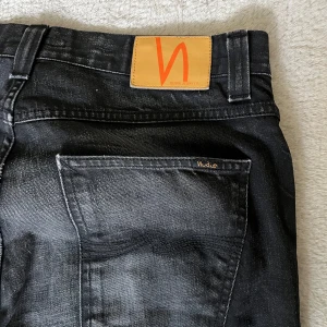 Svarta jeans från Nudie Jeans - Säljer ett par svarta jeans från Nudie Jeans med klassisk femficksdesign och raka ben. Jeansen har subtila slitningar och en snygg läderpatch med logga bak i midjan.