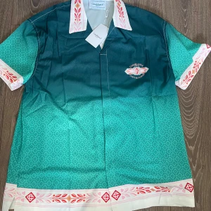 Grön kortärmad skjorta från Casablanca - Säljer en grön kortärmad skjorta från Casablanca med diskret mönster och broderad logga på bröstet. Skjortan har vit krage och ärmslut med röda och rosa detaljer samt knappar framtill. Perfekt för dig som vill sticka ut med stil.