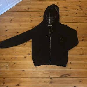 Svart hoodie ifrån Burberry - Svart hoodiejacka från Burberry med dragkedja framtill och klassiskt rutigt foder i huvan och insidan. Jackan har långa ärmar, två fickor fram och justerbar huva med snörning. Perfekt för en stilren och avslappnad look. Priset är inte hugget i sten vid snabb affär!