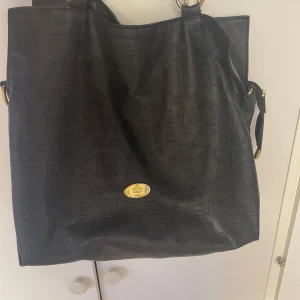 Svart axelväska från Fendi - Säljer en svart axelväska från Fendi med präglad logotyp över hela väskan och guldfärgade metalldetaljer. Väskan har ett rymligt innerfack med klassiskt Fendi-mönstrat tyg och dragkedja. Perfekt för dig som vill ha en lyxig och stilren accessoar.