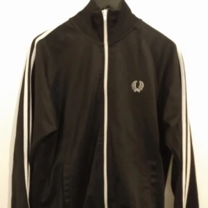 Fred perry tröja - Svart track jacket från Fred Perry med vita ränder längs ärmarna och vit dragkedja framtill. Jackan har hög krage och Fred Perry-logga broderad på bröstet. Perfekt för en sportig och stilren look. Köpte den förra sommaren o använde den bara når gånger 