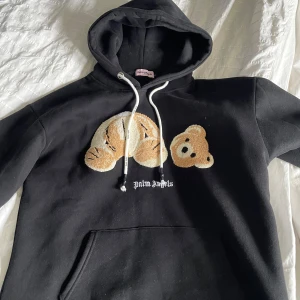 Svart hoodie från Palm Angels med nallemotiv - Svart hoodie från Palm Angels med broderat nallemotiv på bröstet och vita snören i huvan. Stor magficka framtill och klassisk passform. Perfekt för dig som gillar streetwear och unika detaljer. Helt fabriksny