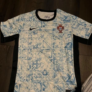 Portugal fotbollströja Nike - Säljer en Portugal fotbollströja från Nike med unikt blått mönster på vit bas. Tröjan har korta ärmar, svarta sidopaneler och Portugal-emblem på bröstet. Perfekt för dig som vill sticka ut på planen eller heja på landslaget.