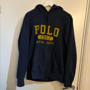 Mörkblå hoodie från Ralph Lauren - Säljer en snygg mörkblå hoodie från Ralph Lauren med gul text 'POLO 1967 ATHL DEPT' på framsidan. Riktigt skön hoddie är skön till sommaren